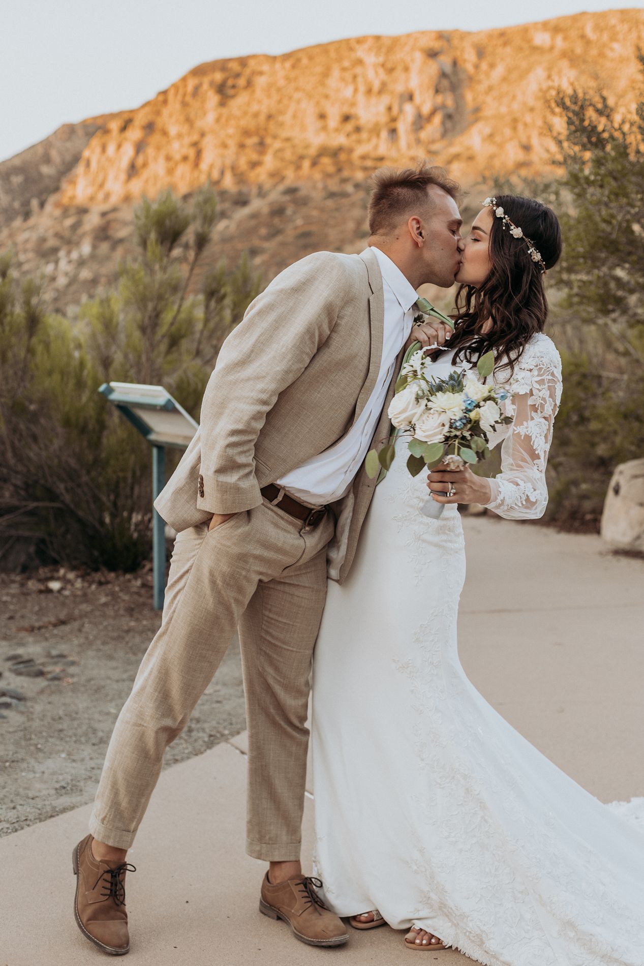 Bride and groom sharing a romantic kiss outdoors, with a scenic mountain backdrop, bride holding a white bouquet.