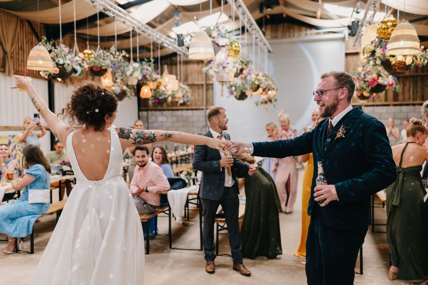 Bride and groom share first dance in rustic barn venue adorned with hanging floral arrangements and fairy lights.