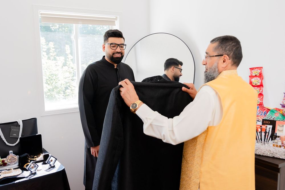 Two men in traditional South Asian attire, one being fitted with a black sherwani in a bright room with a mirror.