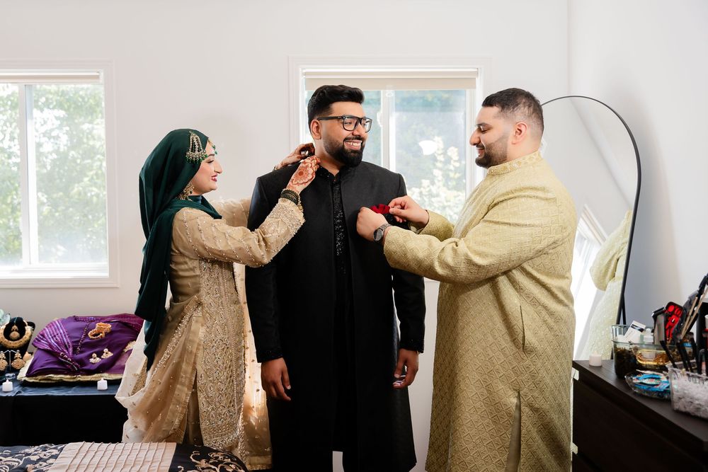 South Asian wedding party helps groom dress in traditional black sherwani attire indoors.