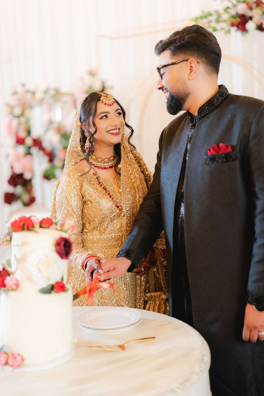 South Asian bride and groom in traditional attire cutting wedding cake, bride in gold lehenga, groom in black sherwani.