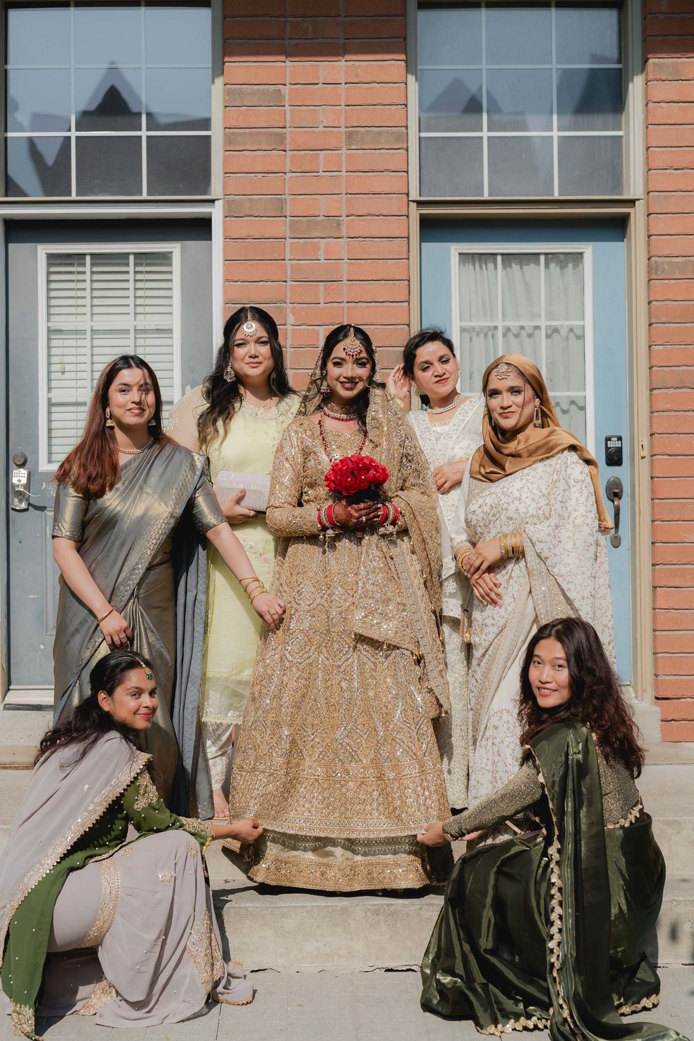 Indian bride in gold lehenga holds red bouquet surrounded by bridesmaids in traditional attire outside brick building.
