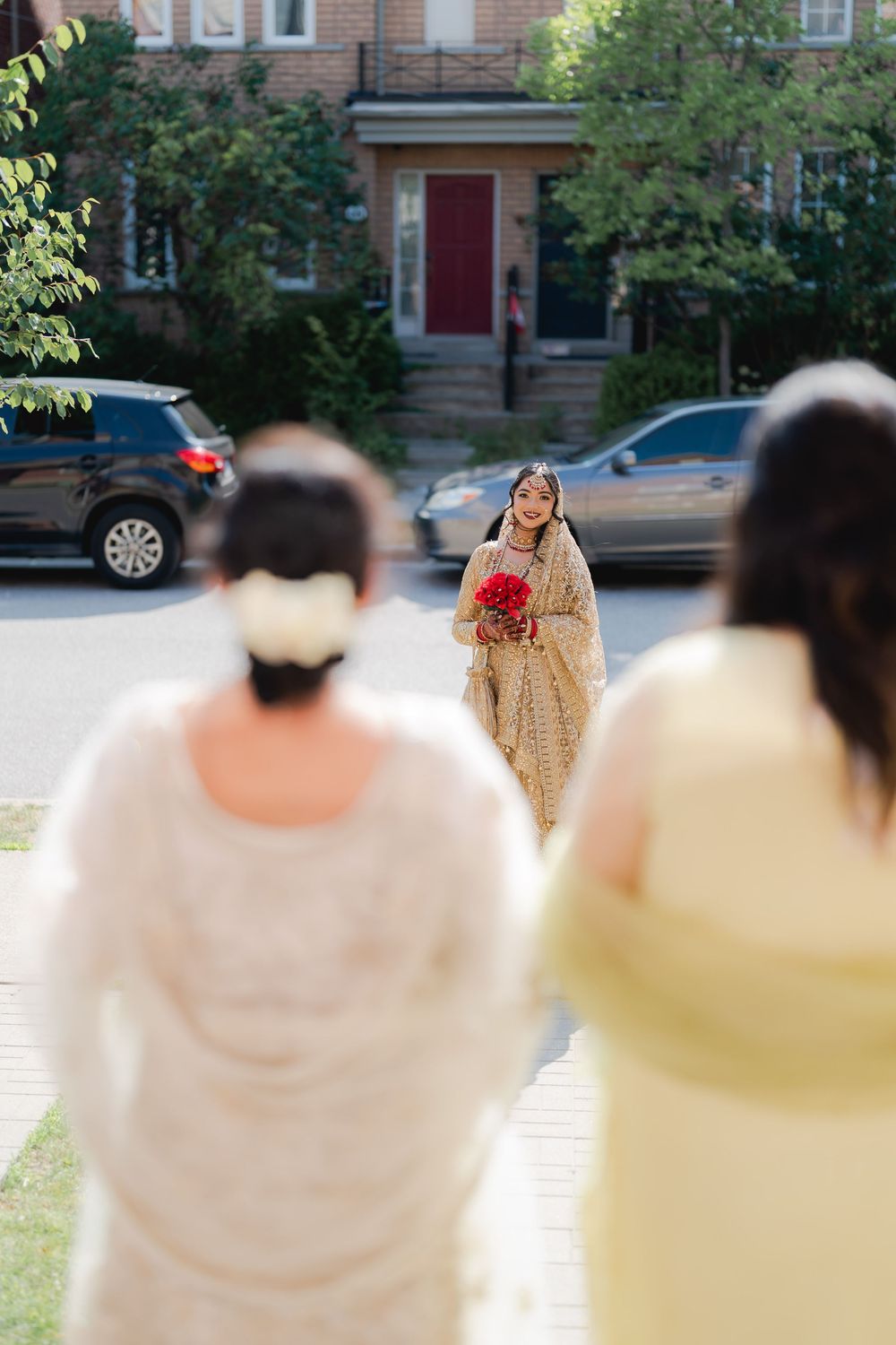 Bride in gold lehenga holding red bouquet stands on street while two women in white and yellow watch.