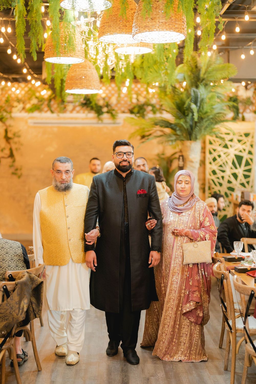 A man in black sherwani walks with elderly parents in traditional South Asian attire at an elegant wedding venue.