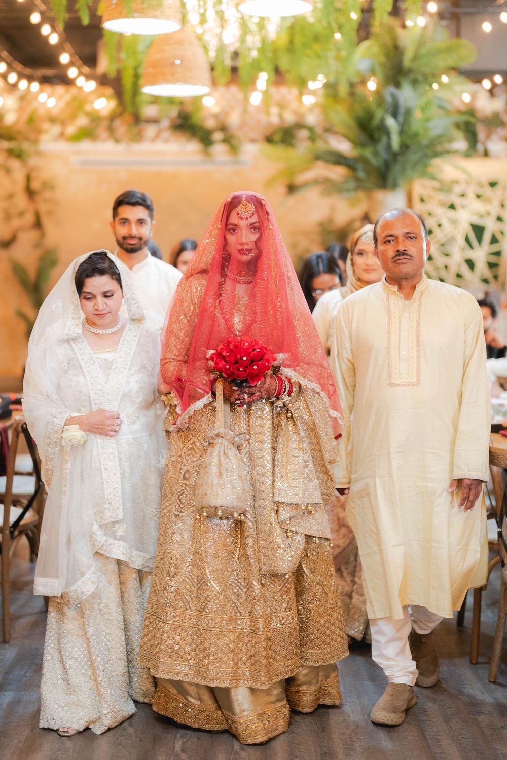 Bride in golden lehenga with red dupatta walks down aisle with family at South Asian wedding ceremony.