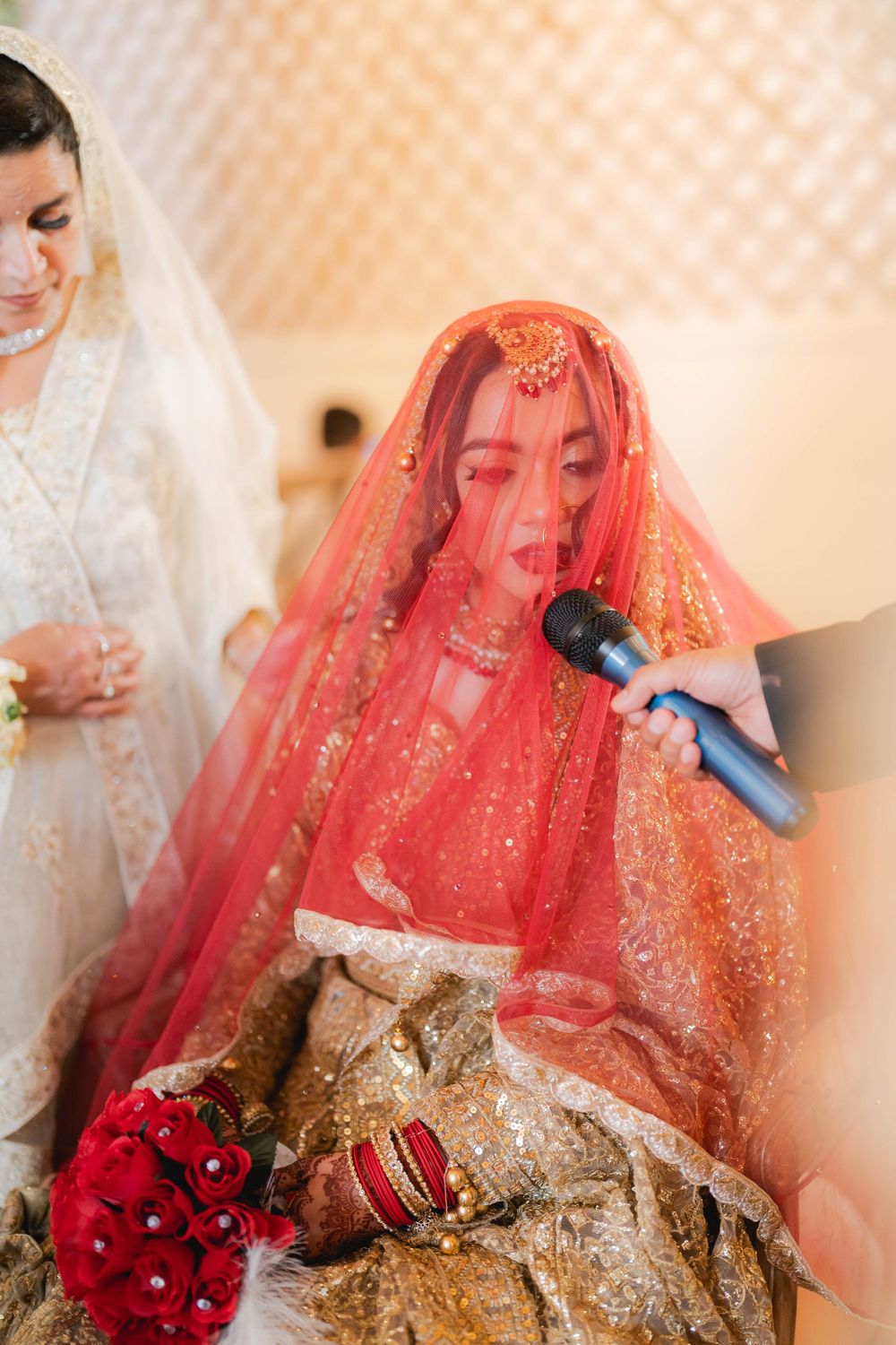 Bride in red dupatta and gold lehenga speaks into microphone at South Asian wedding ceremony.