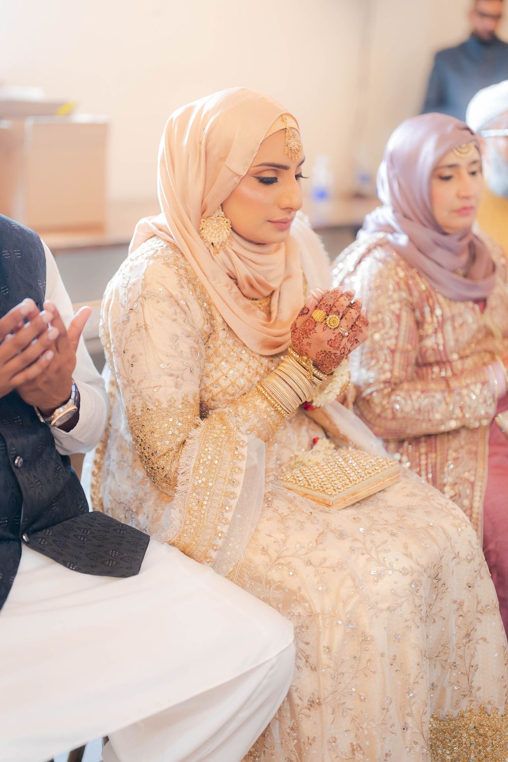 Muslim bride in cream hijab and embroidered lehenga with mehndi hands prays at wedding ceremony.