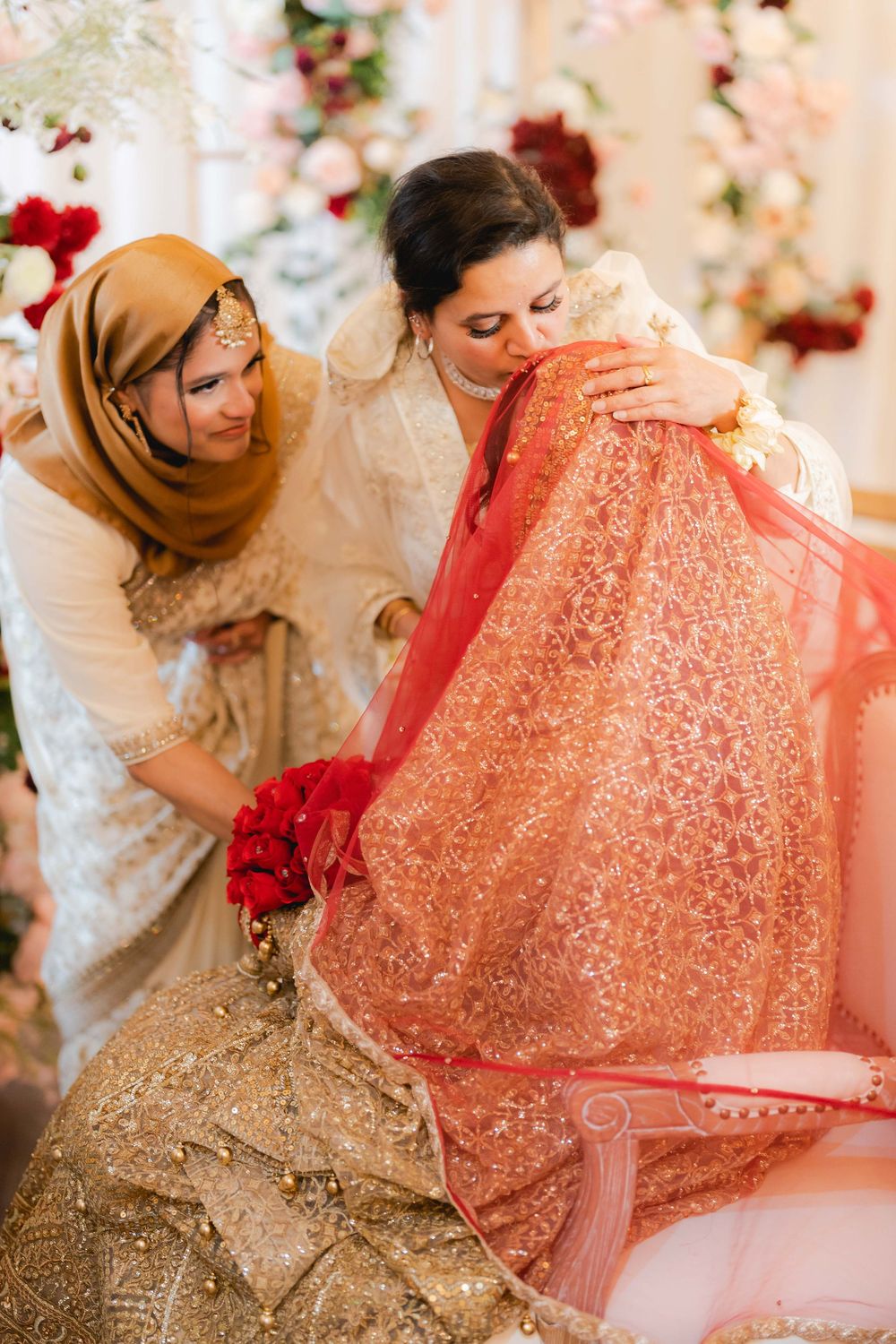 Woman in hijab adjusts bride's red dupatta during South Asian wedding ceremony with floral backdrop.