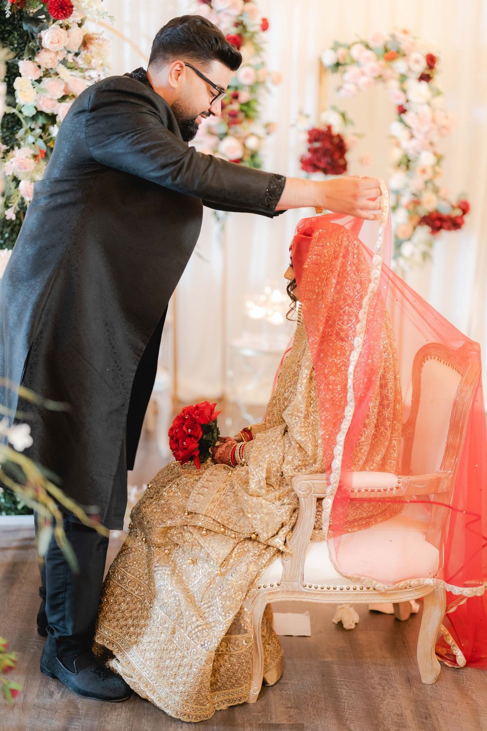 Groom in black kurta lifting red dupatta over bride in gold lehenga seated on white chair at Indian wedding ceremony.
