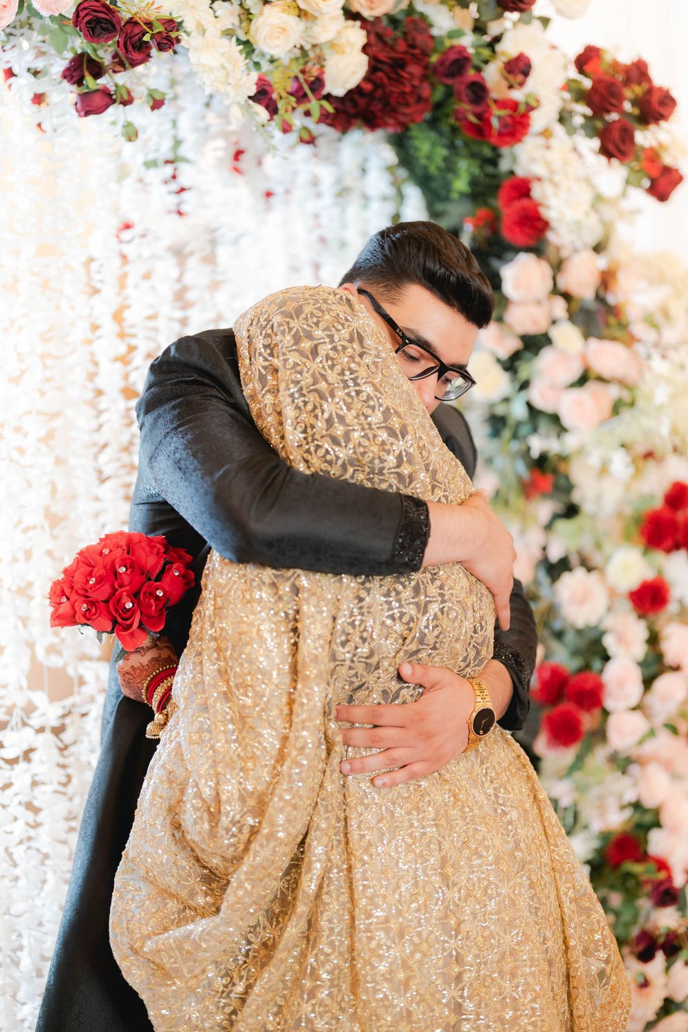 Couple embracing in golden bridal outfit amid red and white floral backdrop at South Asian wedding ceremony.