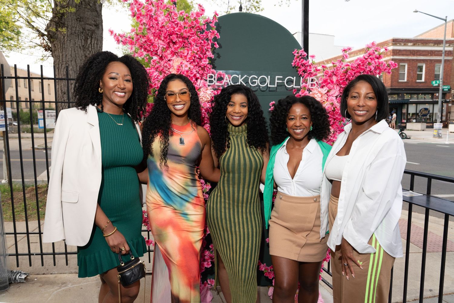 Six women posing together outdoors at a Black Golf Club event, surrounded by pink floral decorations.