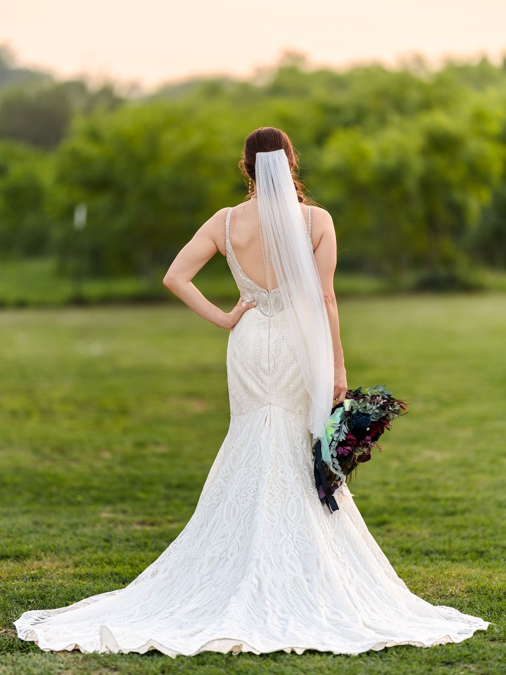 An elegant outdoor bridal portrait from behind, showcasing the intricate back detail of the wedding gown and a long veil in a grassy field.