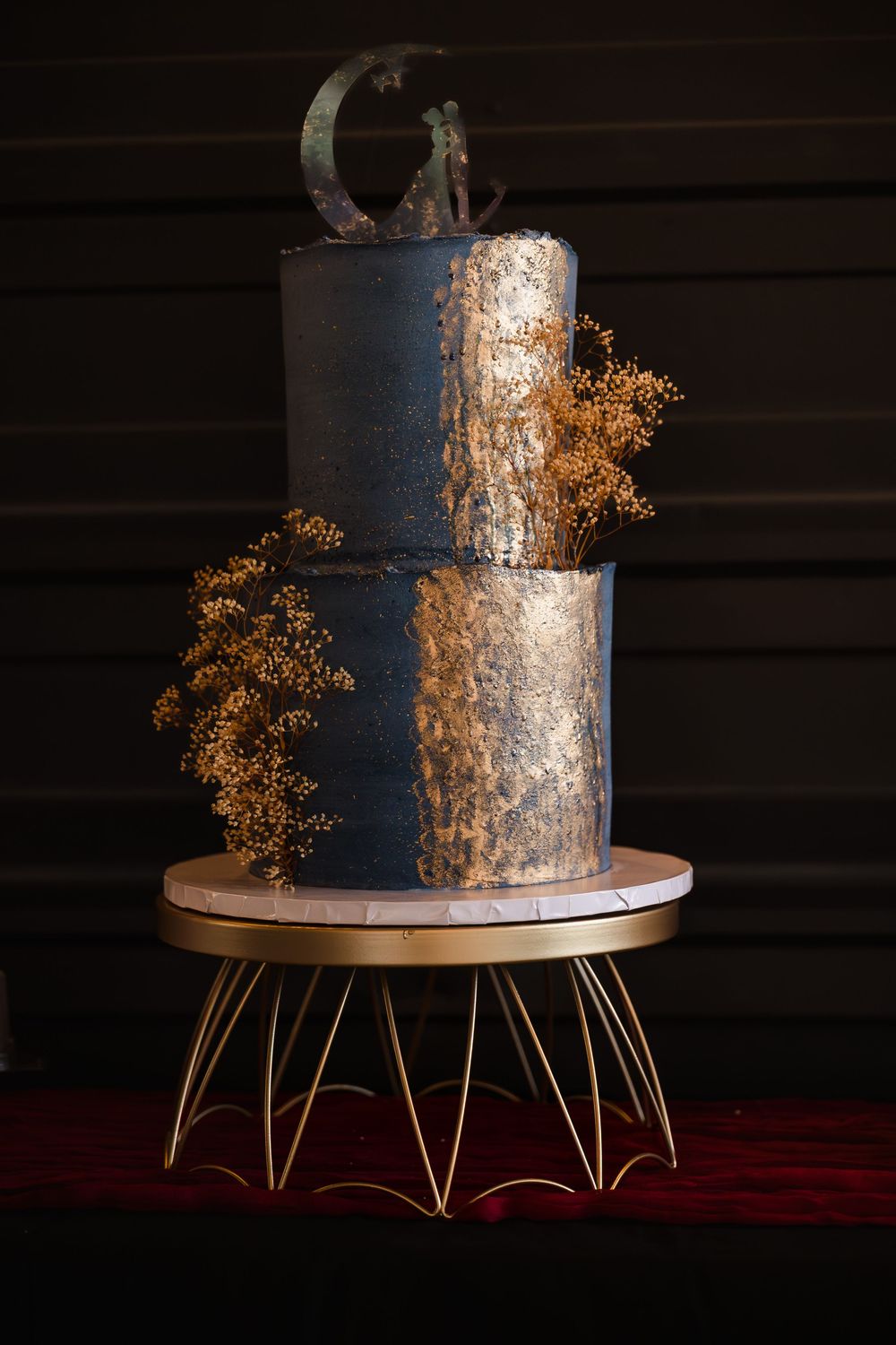 Editorial shot of a modern three-tier dark wedding cake with gold leaf detailing on a decorative gold stand.