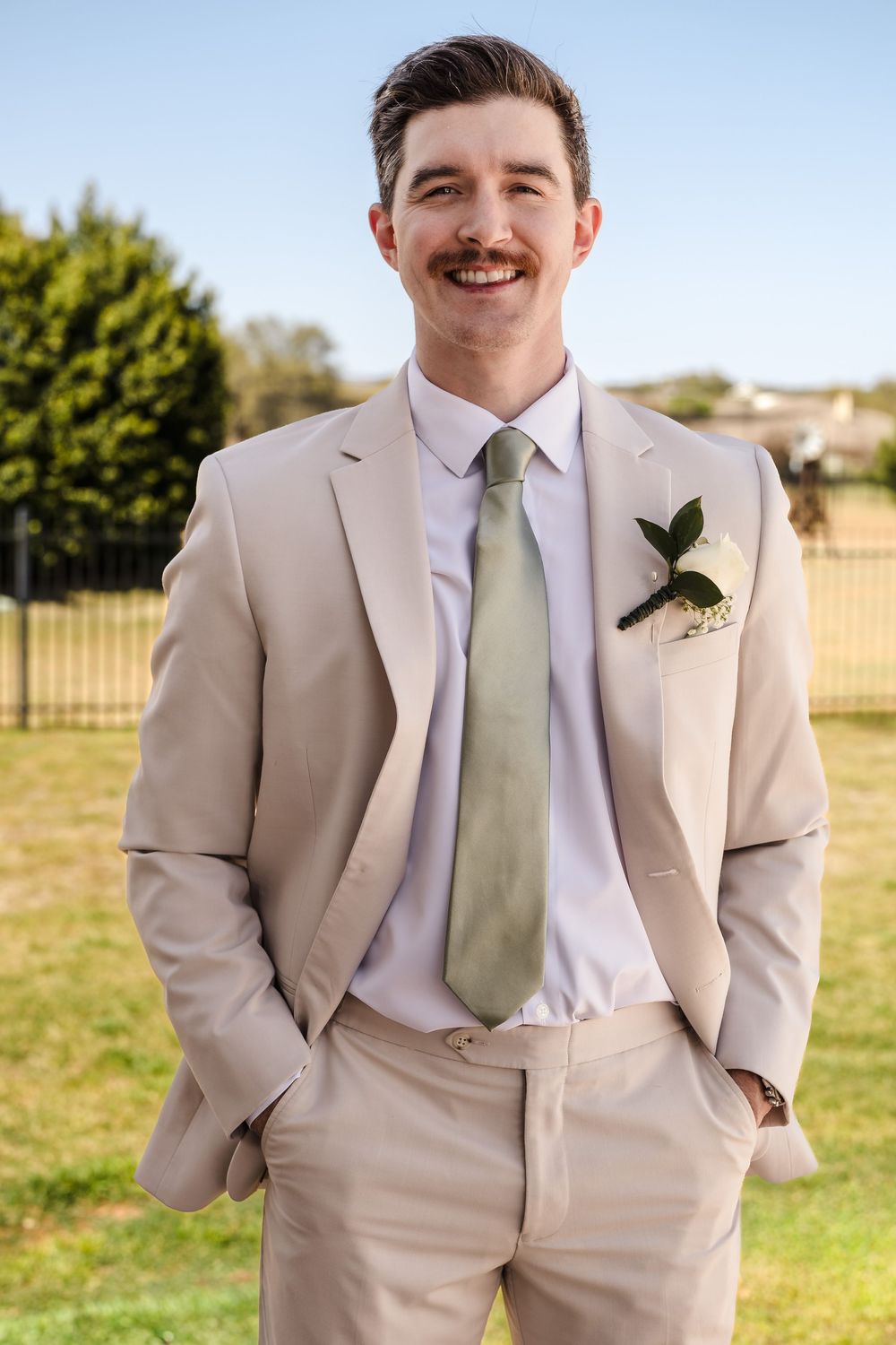 A classic outdoor lifestyle portrait of a smiling groom in a tan suit, captured in soft natural light.