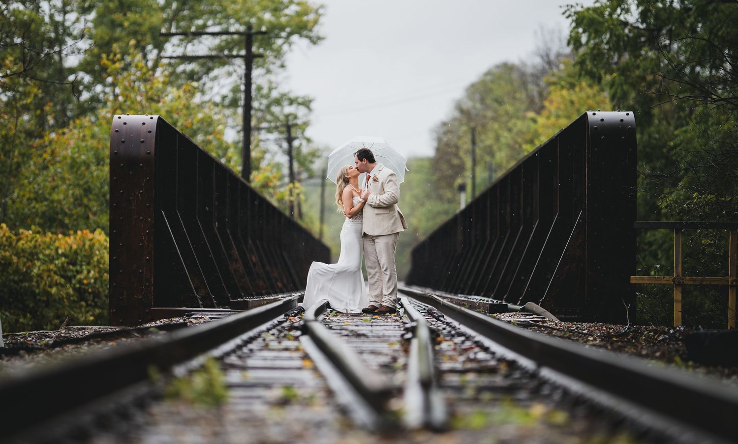 Bride and groom sharing a romantic kiss on railroad tracks on a bridge surrounded by lush green trees.