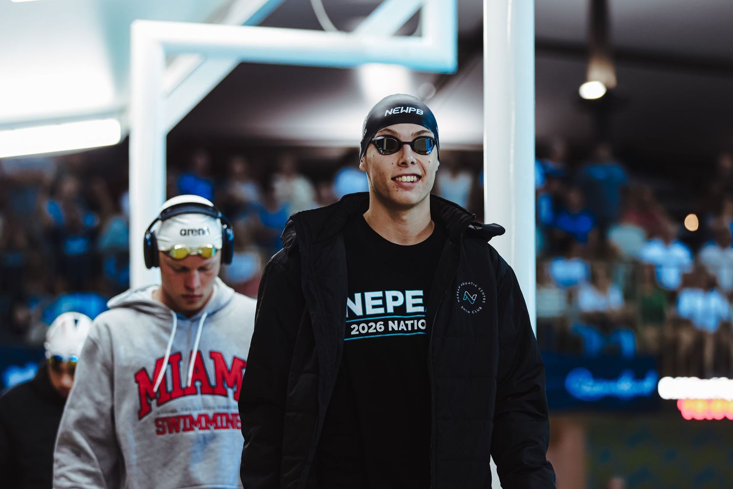 Swimmer in black NEPE 2026 jacket and cap walks poolside at a competitive swim meet with Miami Swimming athlete behind.