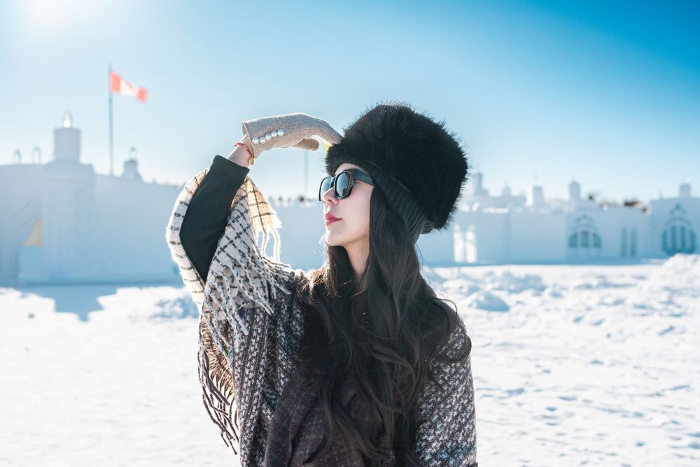 Woman in fur hat and sunglasses posing in snowy winter landscape under bright blue sky.