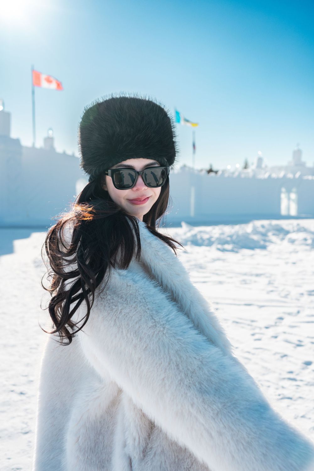 Smiling woman in white fur coat and black fur hat stands before snowy ice castle with Canadian flag in background.