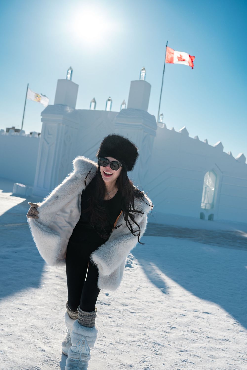 Woman in white fur coat and black hat poses playfully in snowy Canada with ice castle and Canadian flag backdrop.