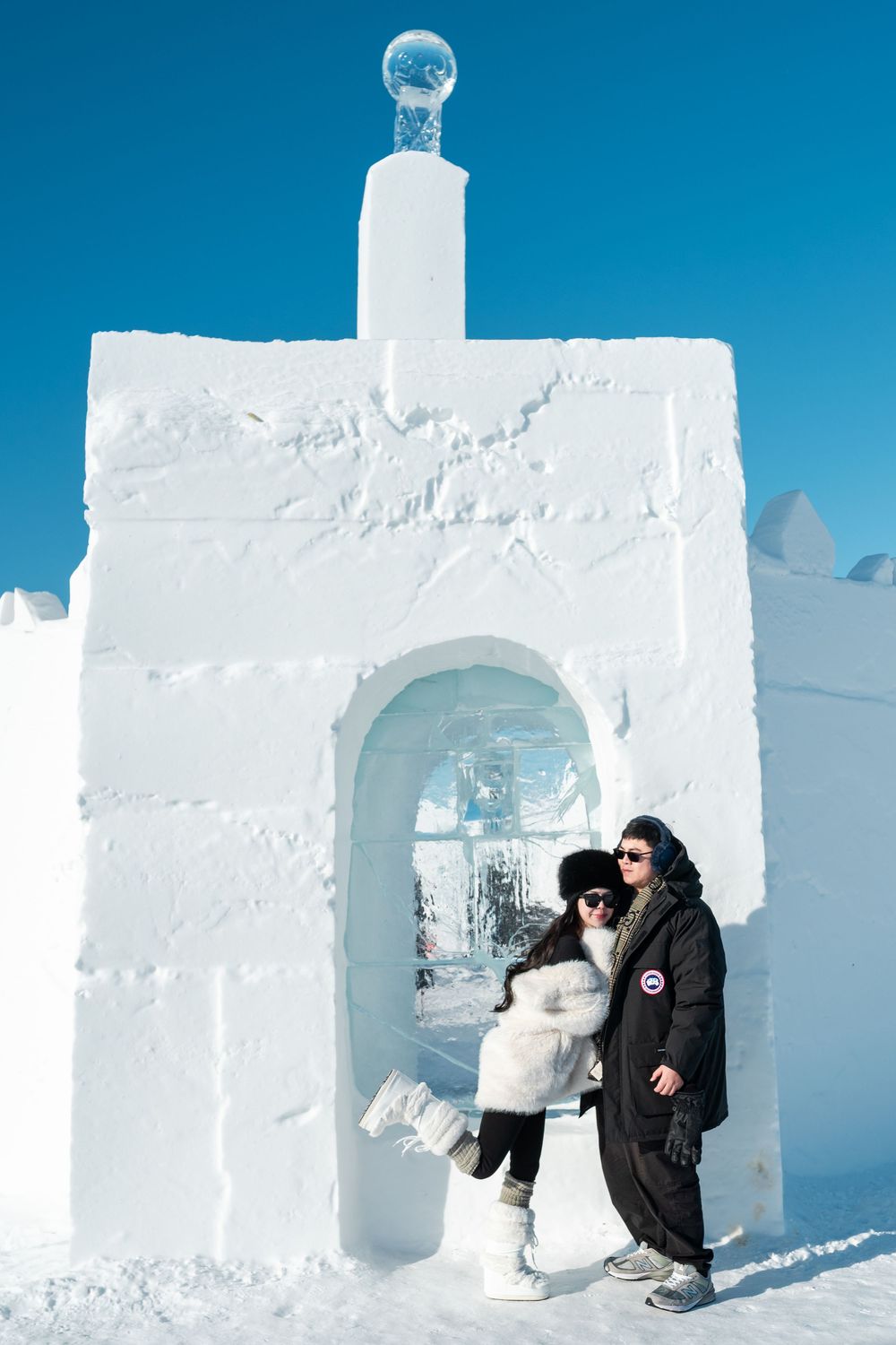 Couple posing in front of a white snow and ice sculpture gateway under a bright blue winter sky.