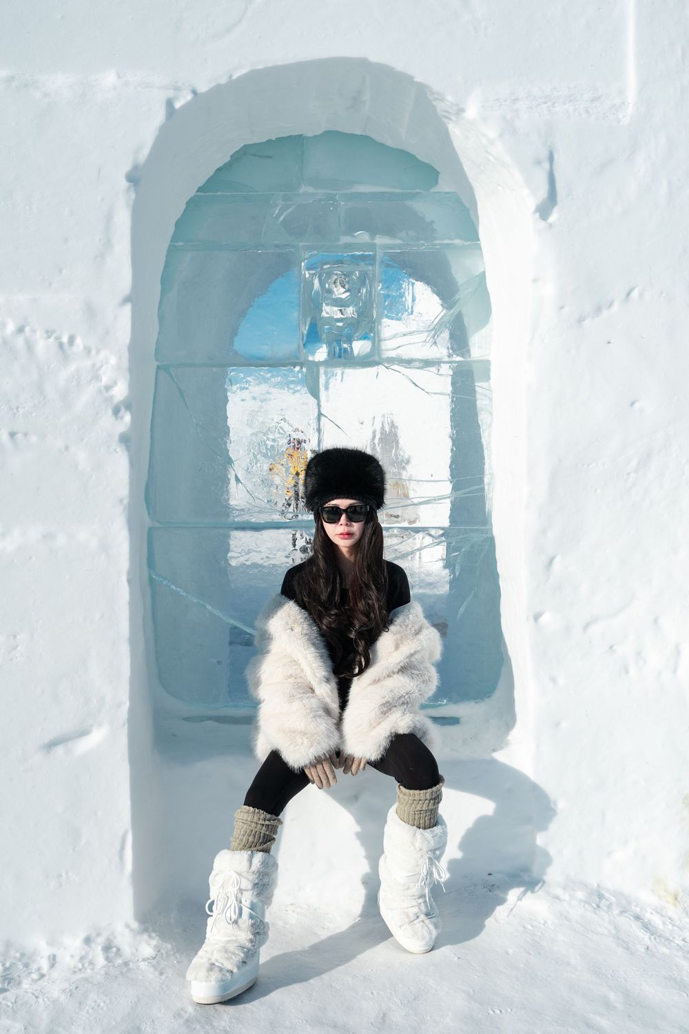 Woman in fur coat and black hat sitting in an arched ice window of a snow castle on a sunny day.