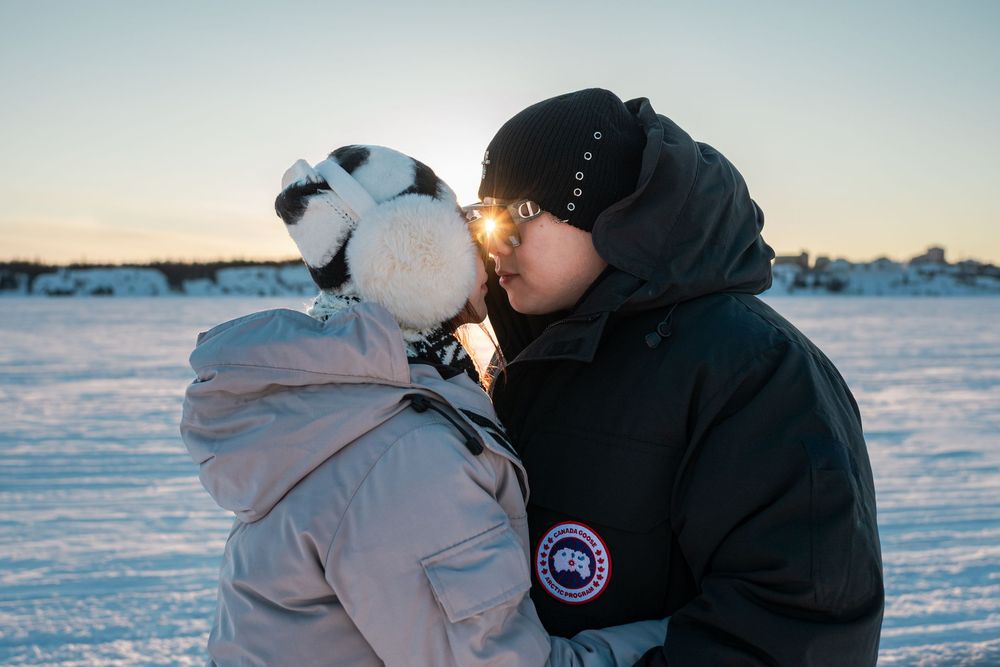 Two people sharing a kiss outdoors in snowy winter landscape wearing Canada Goose jackets at sunset.