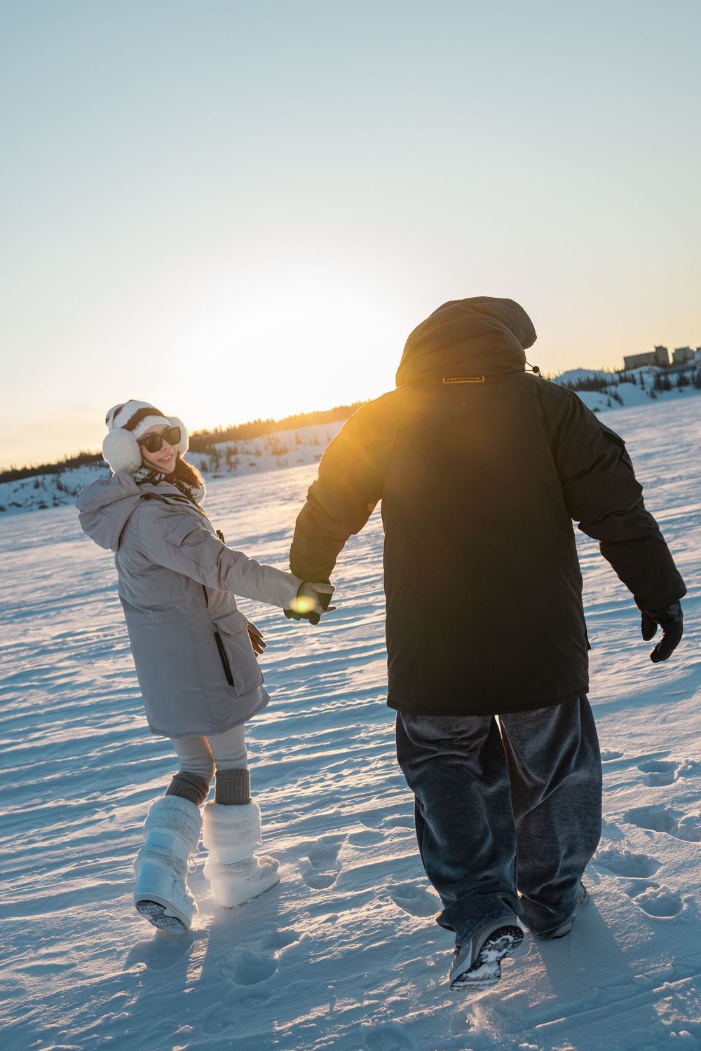 Couple holding hands walking across a snowy field at golden sunset in winter.