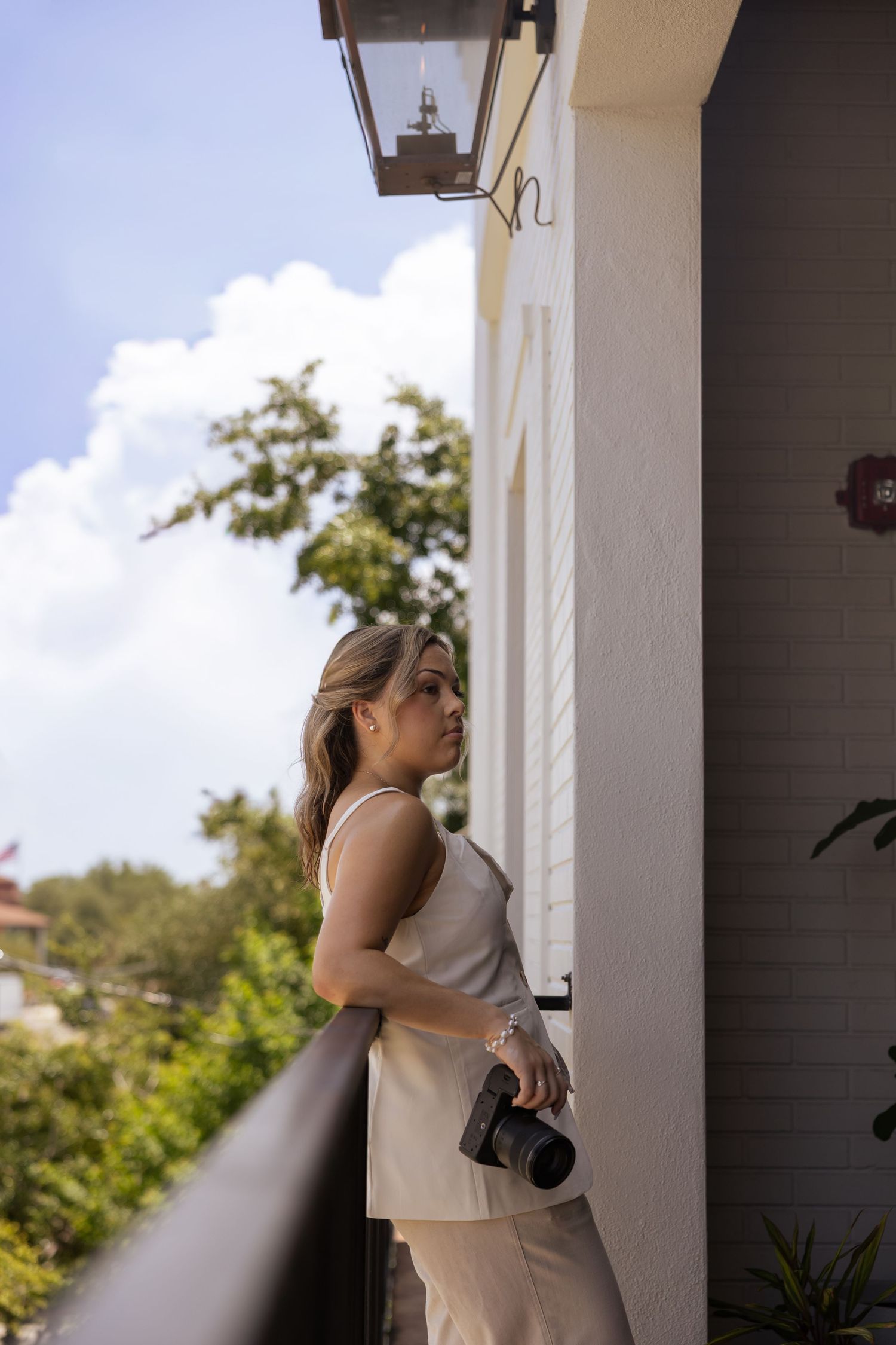 Woman in a cream sleeveless top and trousers leans on a balcony railing holding a camera beneath a lantern, with trees and blue sky behind.