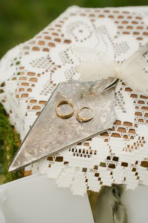 Two gold wedding rings resting on a decorative silver tray atop elegant white lace fabric at an outdoor ceremony.