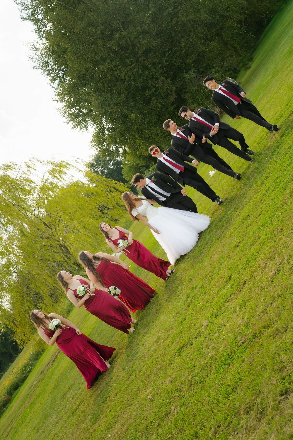 Wedding party walks outdoors on green grass, bride in white gown, bridesmaids in red dresses, groomsmen in black suits.