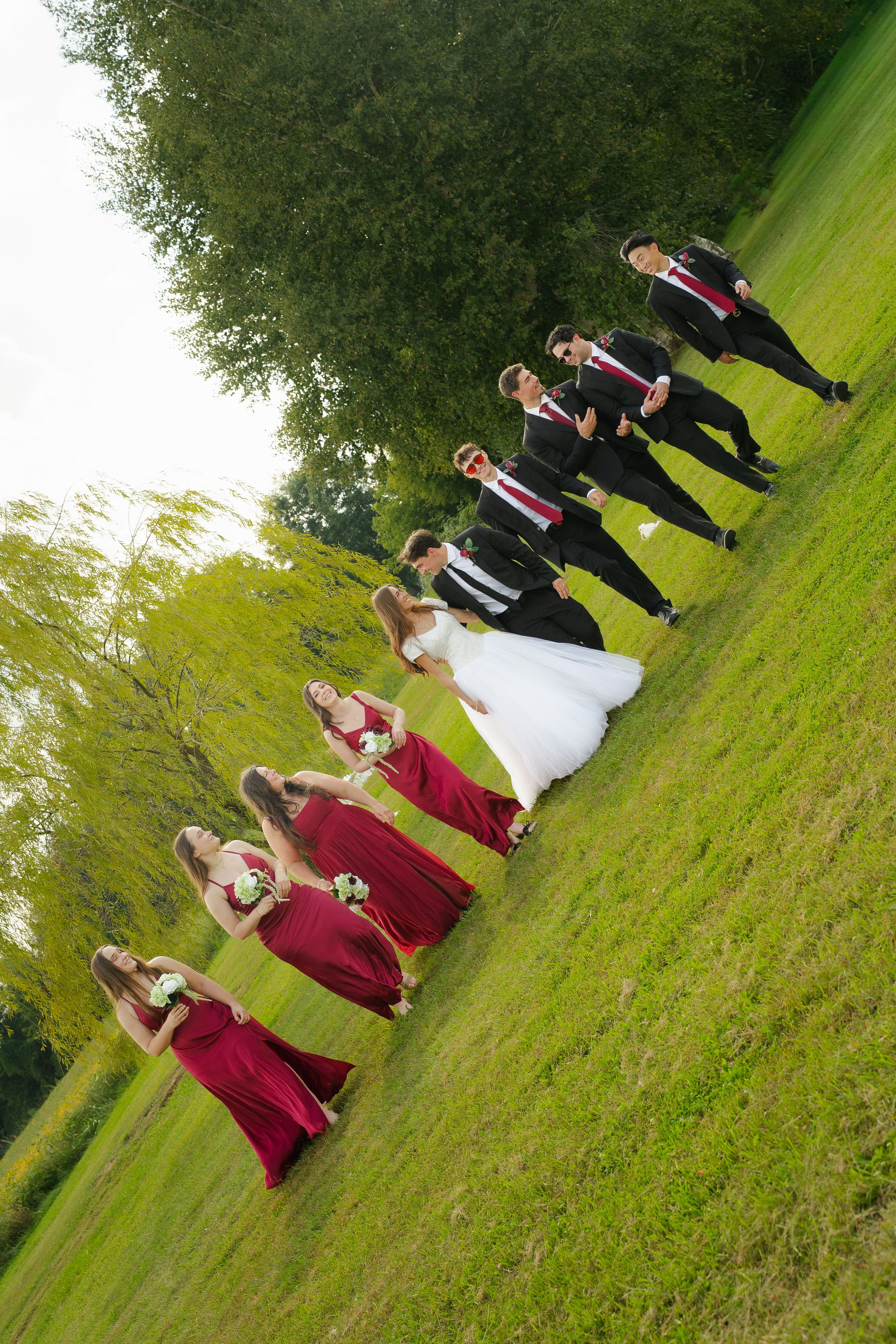 Wedding party walks outdoors on green grass, bride in white gown, bridesmaids in red dresses, groomsmen in black suits.
