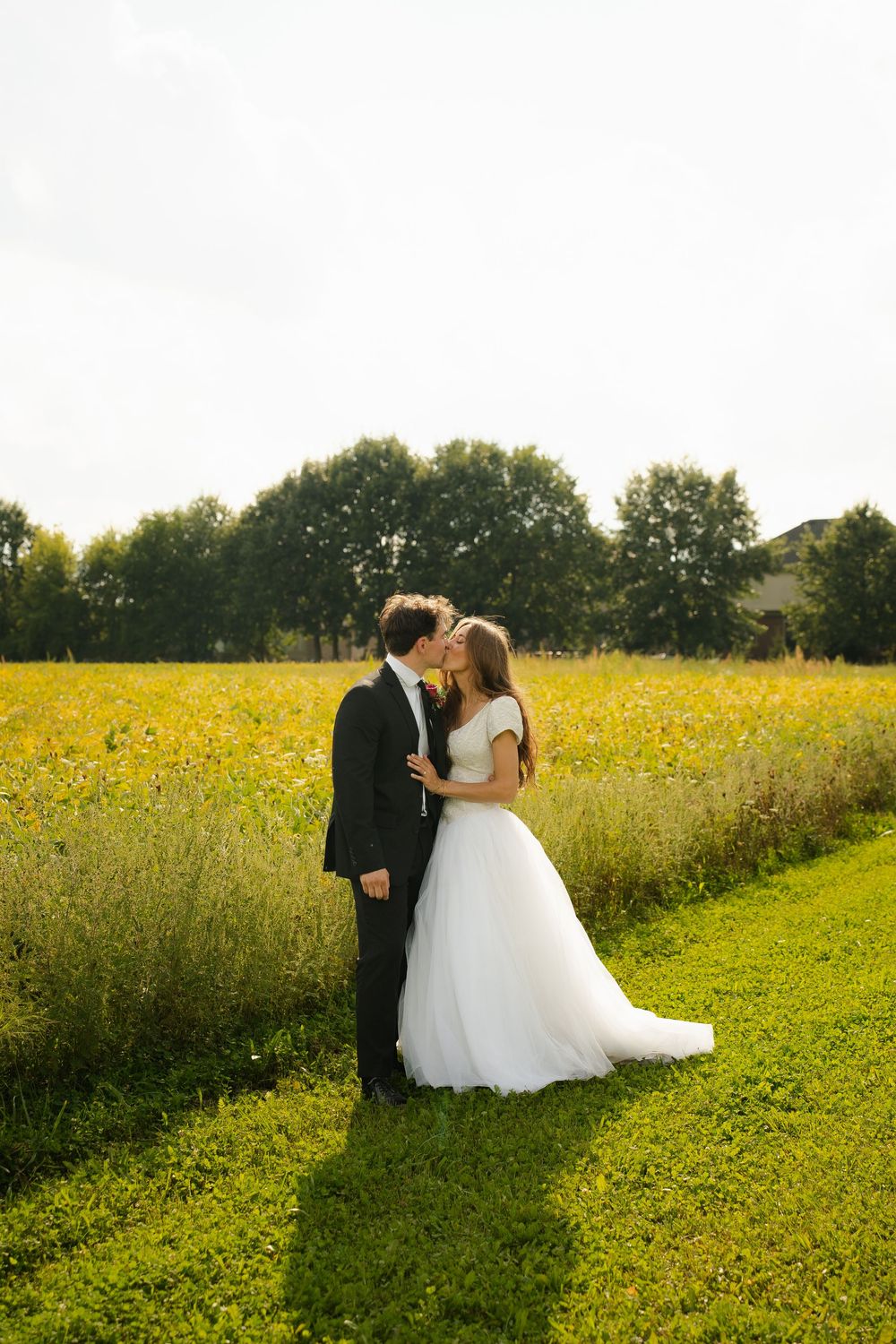 A bride and groom share a romantic kiss in a lush green field with yellow wildflowers and trees in the background.