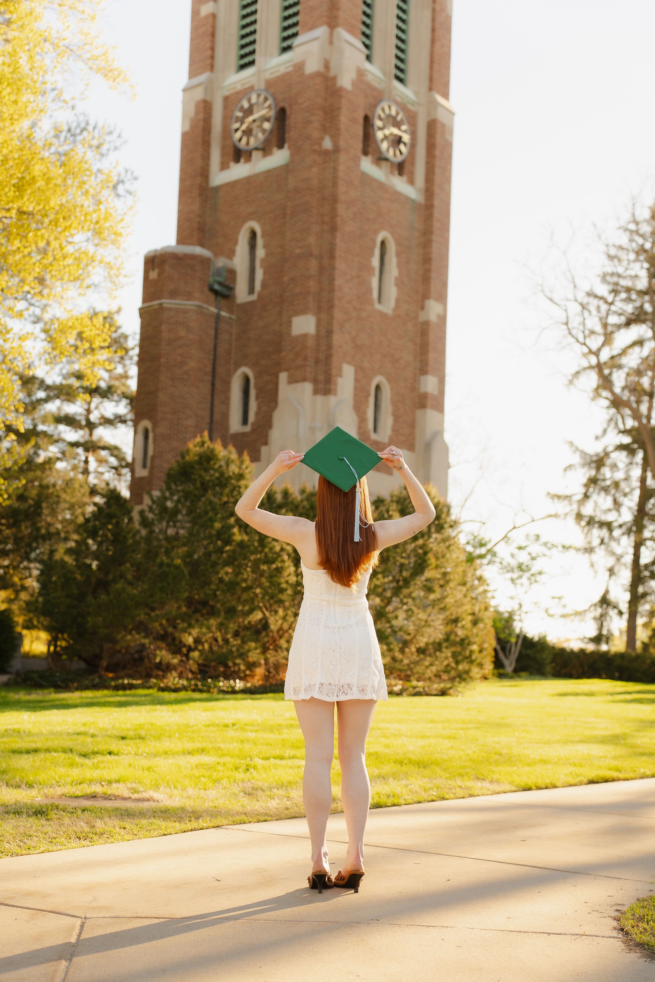 Young woman in white dress holding green graduation cap in front of brick clock tower on sunny campus day.