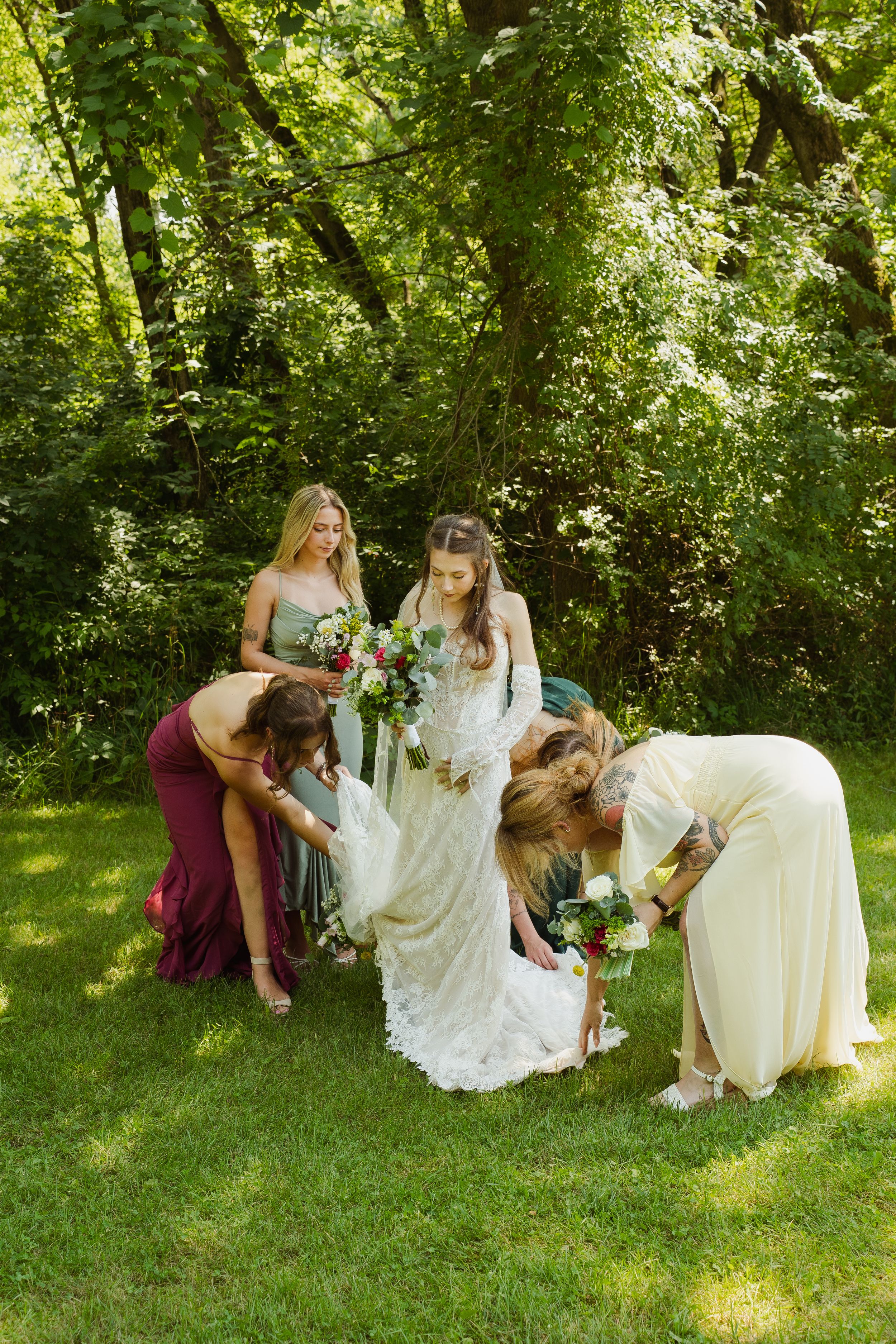 Bridesmaids helping bride with dress outdoors in lush green garden setting during wedding day preparations.
