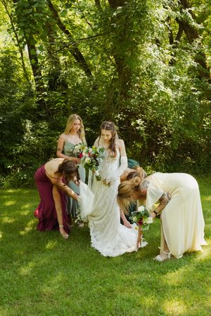 Bridesmaids helping bride with dress outdoors in lush green garden setting during wedding day preparations.