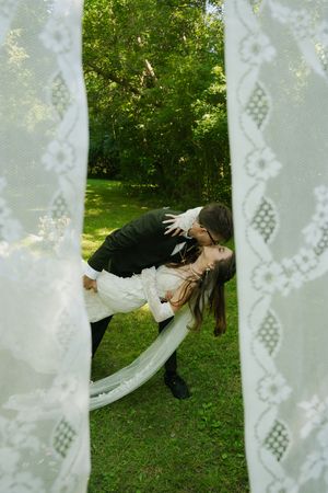 Groom dipping bride for a romantic kiss in lush green garden, framed by elegant white lace curtains.