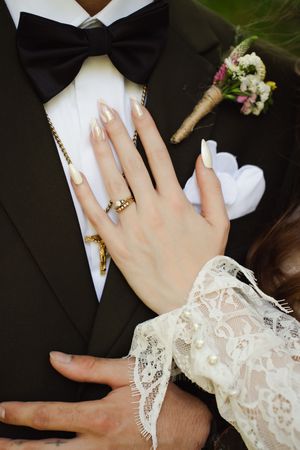 Bride's hand with gold claddagh ring and nude almond nails resting on groom's black tuxedo with boutonniere.