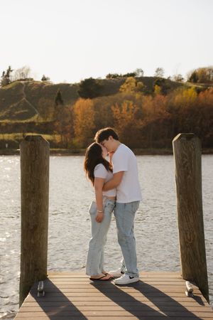 Couple sharing a romantic moment on a wooden dock by a lake with autumn foliage in the background.
