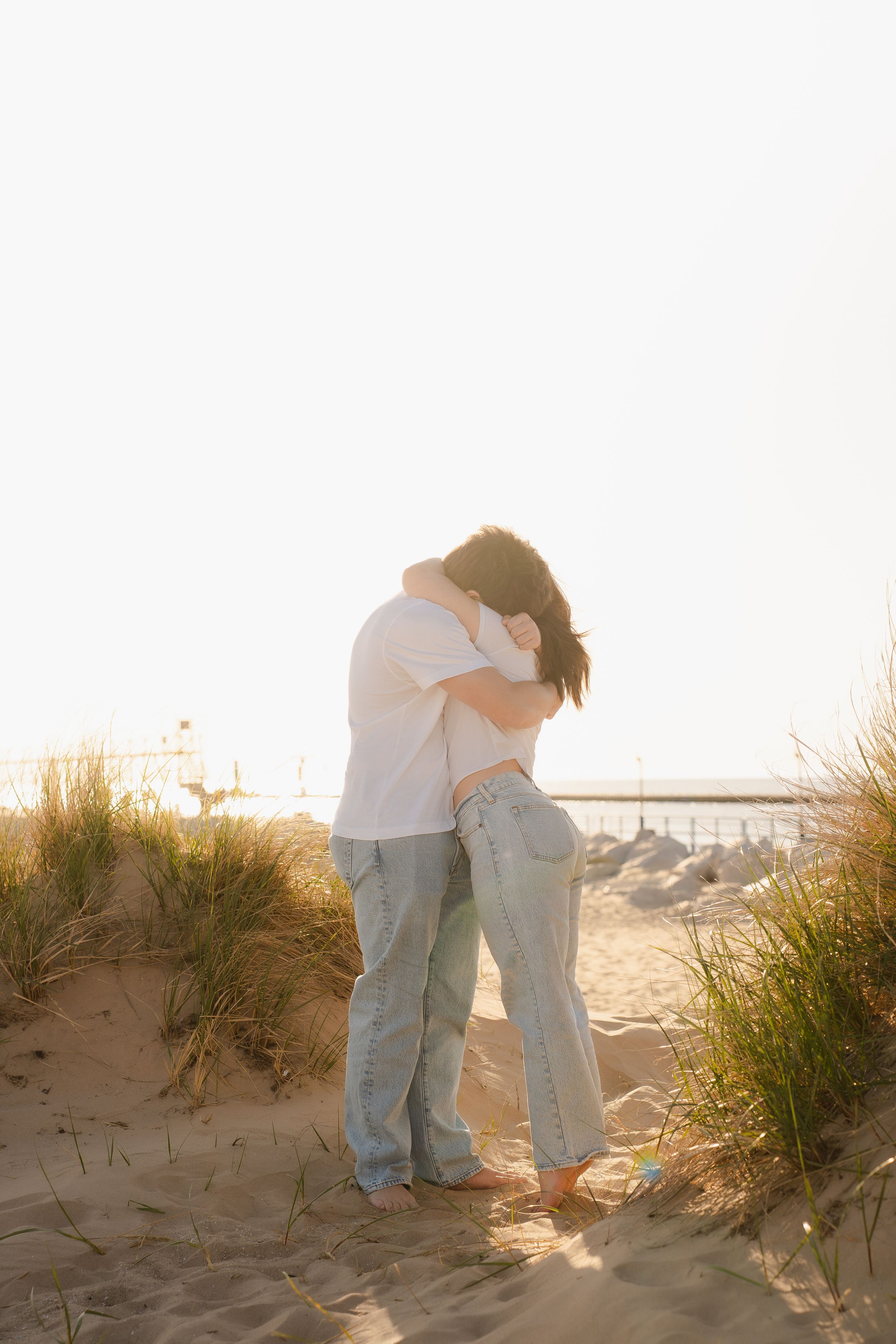 Couple embracing on sandy beach dunes at golden hour, wearing jeans and casual tops, with ocean in background.