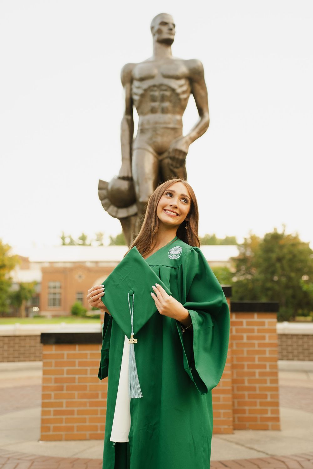 Smiling graduate in green cap and gown holds mortarboard before a large Spartan statue at Michigan State University.