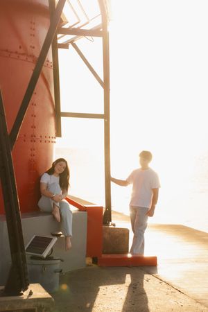 Young couple near red industrial structure at golden hour, woman sitting while man stands nearby.