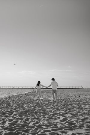 Black and white photo of a couple holding hands on a sandy beach with a pier in the background.