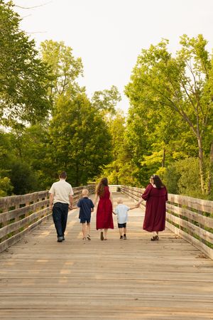 Family of five walking hand-in-hand across a wooden bridge surrounded by lush green trees during golden hour.