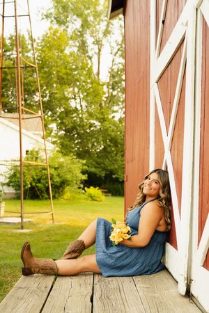 Young woman in blue dress and cowboy boots sits against red barn holding yellow flowers on wooden deck.