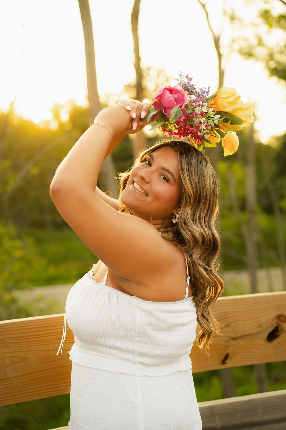 Young woman in white dress wearing colorful flower crown, smiling outdoors at golden hour near wooden fence.