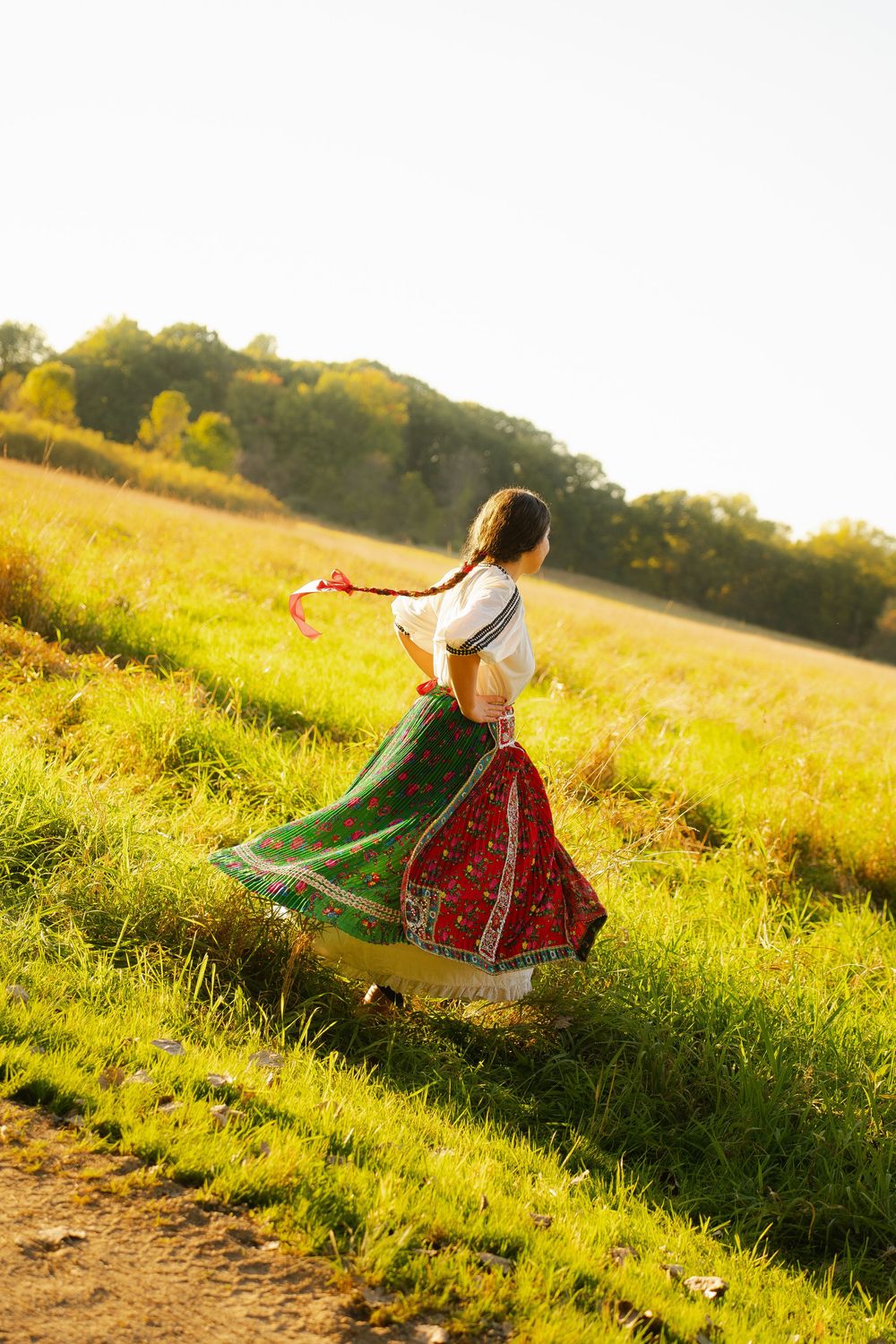 Woman in traditional red and green folk costume twirling in a sunny green meadow with trees in background.