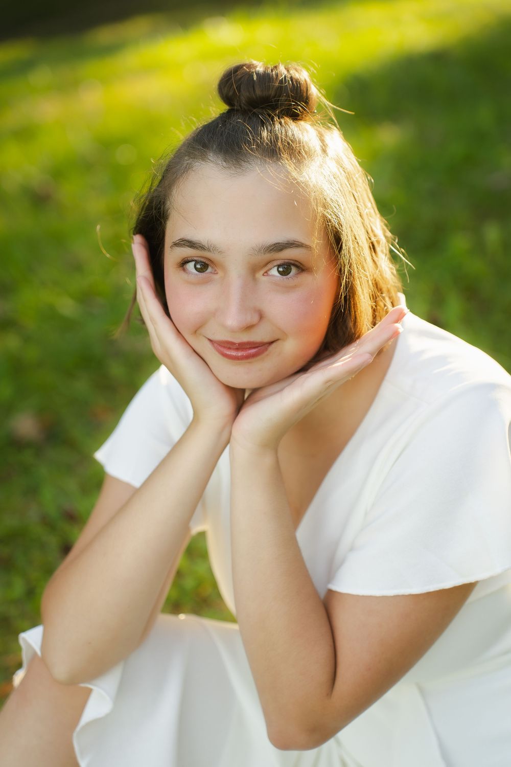 Young woman in white dress sitting on grass, hands framing her smiling face, with a hair bun in sunny park.