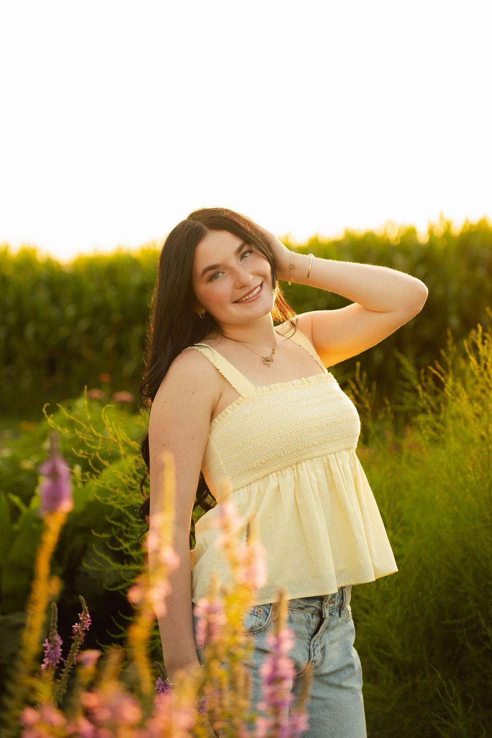 Young woman in yellow top and jeans smiling in a sunlit field with wildflowers during golden hour.