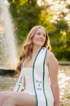 Smiling college student wearing a Delta Tau Class of 2026 graduation sash, seated near a fountain outdoors.