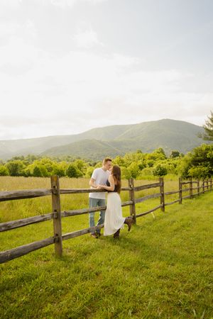 Couple embracing by a rustic wooden fence in a lush green meadow with rolling mountain views in the background.
